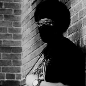 Shaw in front of a brick wall, looking into the camera. He wears an Afro, glasses and a mask that covers his mouth and nose.
