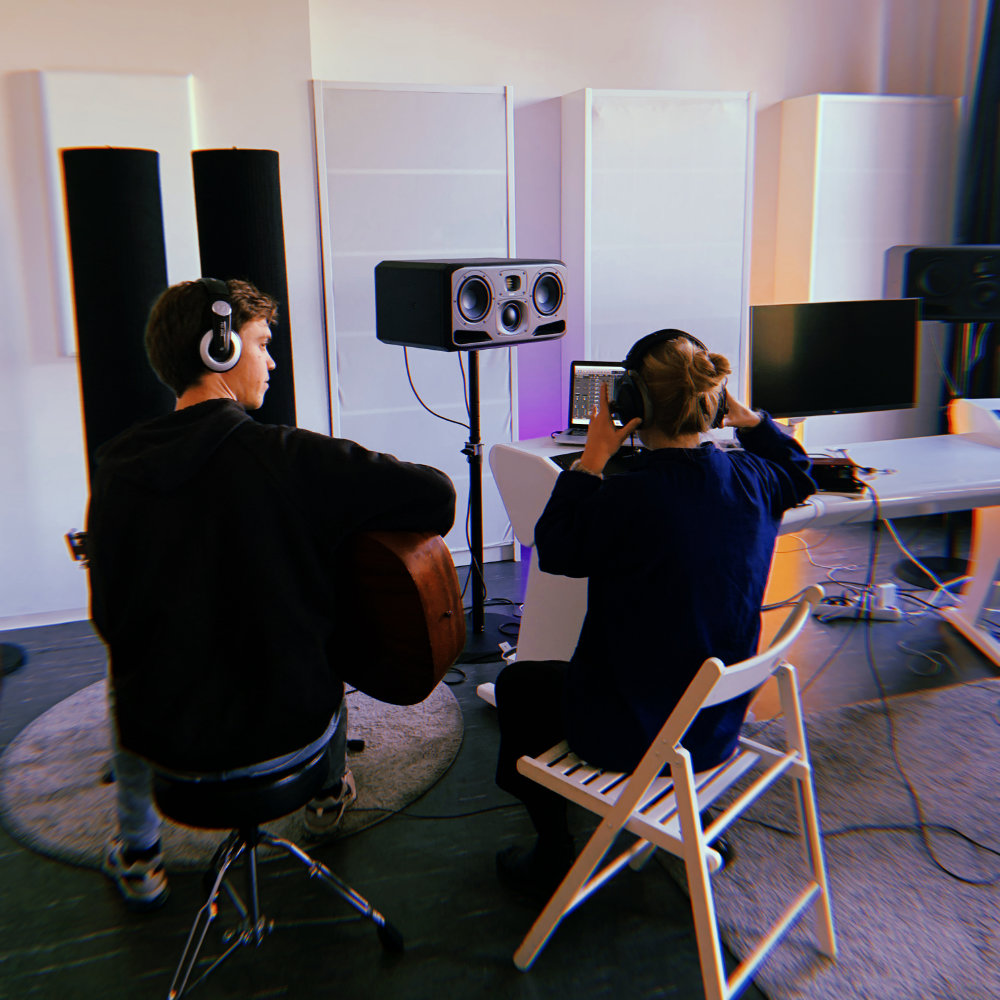 Zwei Teilnehmer*innen des Two participants of the camp writing music together. One playing the guitar, the other is looking at a screen. Both wear headphones.